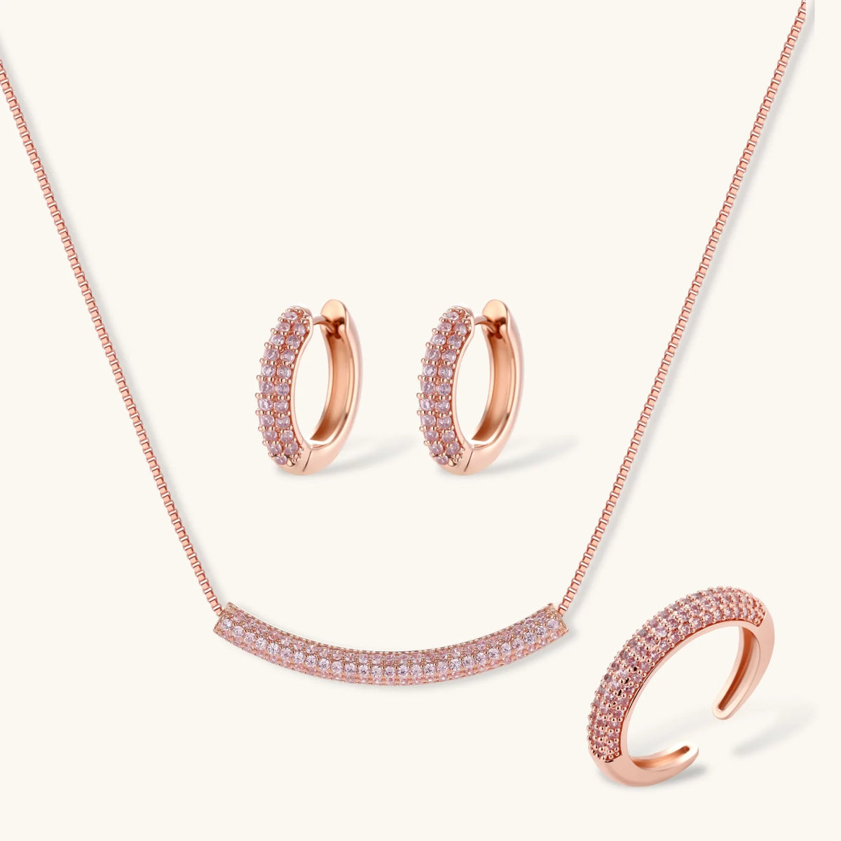 Rose gold jewelry set with pavé cubic zirconia stones including curved necklace, hoop earrings, and adjustable ring on cream background.